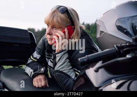 Müde Bikerin mittleren Alters spricht am Herbsttag auf der Straße über ihr schwarzes Motorrad. Hobby - Motorradtourismus. Weibliche Bikerin. Komm Stockfoto