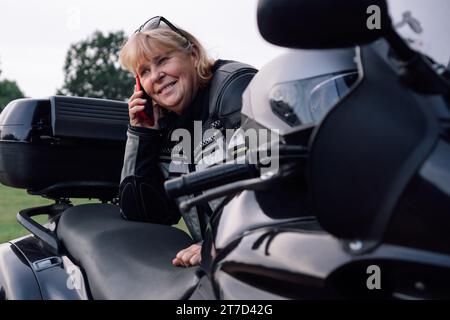 Mobile Kommunikation. Charmante lächelnde Bikerin mittleren Alters, die am Telefon spricht und am Herbsttag auf der Straße auf ihrem Motorrad sitzt. Hobby – Motorrad t Stockfoto