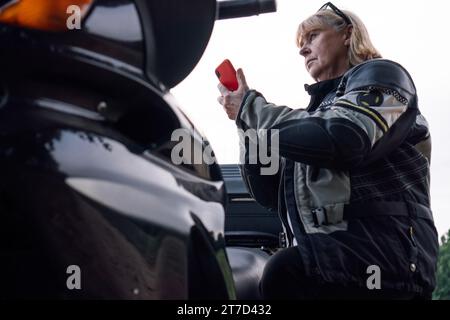 Unteransicht eines selbstbewussten, seriösen, blonden Bikers, der auf rotem Telefon in der Nähe eines schwarzen Motorrads spricht. Hobby - Motorradtourismus. Weibliche Bikerin. Kommunizieren Stockfoto