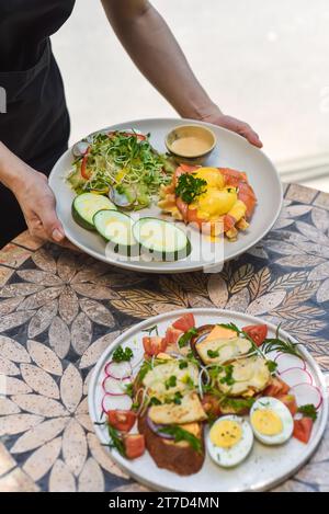 Kellnerin hält einen Teller mit Eiern, bennedict-Waffeln mit geräuchertem Lachs und serviert auf einem Tisch mit Hähnchentoast im Restaurant Stockfoto