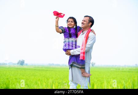 Weite Aufnahme eines glücklichen Dorfmädchens, das mit Flugzeugspielzeug spielt, während der Vater in der Nähe von Farmland hält oder trägt - Konzept der Beziehung, elterlich Stockfoto