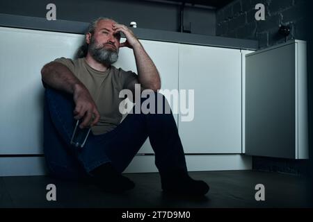 Reifer bärtiger Mann in Depression, der allein auf dem Boden in der Küche sitzt Stockfoto