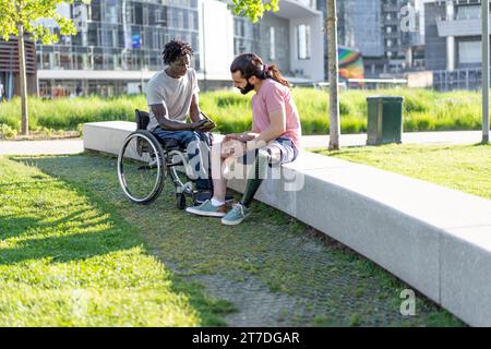 Im Park unterhalten sich zwei Männer mit Inklusivität, einer auf einem Rollstuhl und der andere mit einer Beinprothese. Stockfoto