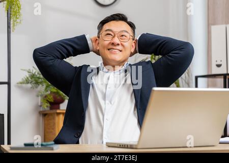 Asiatischer reifer Geschäftsmann Freiberufler macht Pause, lehnt sich nach der Arbeit auf den Stuhl und feiert den Erfolg am Arbeitsplatz im Home Office. Männliche Chinesen im formellen Anzug sorgen für entspannende Hände hinter dem Kopf Stockfoto