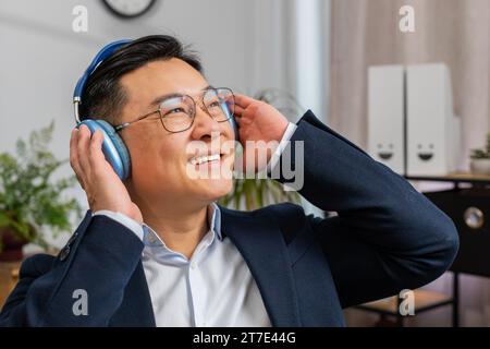 Close-up Porträt glücklicher entspannter asiatischer Geschäftsmann, der im Home-Office an einem Laptop arbeitet und Kopfhörer trägt, der liebste energische Disco-Musik beim Tanzen hört. Freiberuflicher Mann, der sich entspannt, eine Pause macht Stockfoto