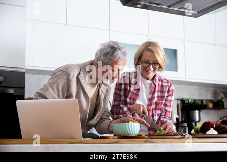 Seniorenpaar kochen gemeinsam in der Küche zum goldenen Hochzeitstag und lesen Rezepte aus dem Internet auf Laptop. Ältere Männer und Frauen machen sich gut Stockfoto