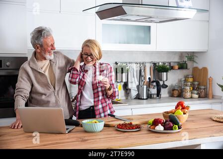 Seniorenpaar kochen gemeinsam in der Küche zum goldenen Hochzeitstag und lesen Rezepte aus dem Internet auf Laptop. Ältere Männer und Frauen machen sich gut Stockfoto