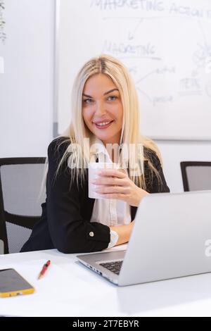 Junge schöne blonde, fröhliche junge Geschäftsfrau lächelt und blickt in die Kamera und trinkt einen Kaffee, während sie im Büro arbeitet Stockfoto