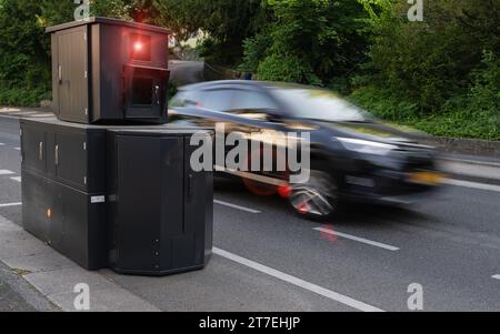 Kamera zur Geschwindigkeitsüberwachung am Straßenrand mit einem Auto in Hochgeschwindigkeitsbewegungsunschärfe, Laubhintergrund Stockfoto