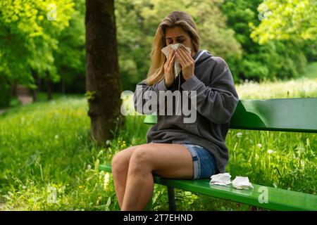 Frau niest auf einer grünen Parkbank, benutzte Taschentücher daneben, wegen einer Pollenallergie Stockfoto