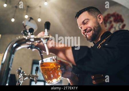 Ein lächelnder, stilvoller Bärenbarmann in schwarzer Uniform mit einer Schürze, der frisches Lagerbier in einen Glasbecher an der Bar zapft. Erfolgreiche Leute, Bierkonzern Stockfoto