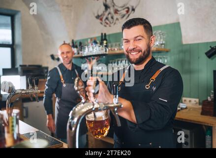 Stilvoller bärtiger Barkeeper in schwarzer Uniform, lächelnd in die Kamera, Bierzapfen an der Bar und Kellner mit Tablett. Erfolgreiche Teamarbeit, Freundlichkeit Stockfoto