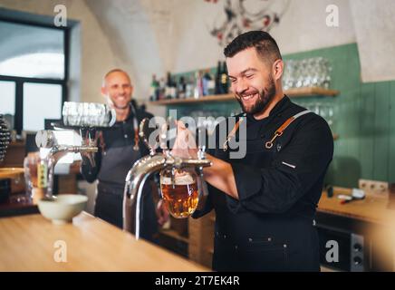 Stilvoller bärtiger Barkeeper in schwarzer Uniform, lächelnd in die Kamera, Bierzapfen an der Bar und Kellner mit Tablett. Erfolgreiche Teamarbeit, Freundlichkeit Stockfoto