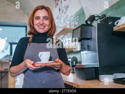 Fröhlich fröhliche lächelnde Kellnerin in der Nähe der Kaffeemaschine, die in einem kleinen Café aromatischen Espresso mit weißer Tasse trug. Glückliche Menschen, Kaffeekonsum, bev Stockfoto