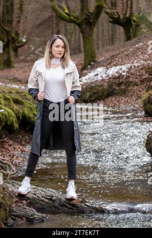 Frau in einem beigefarbenen Mantel, die auf einem Felsen in einem Bach in einem verschneiten Wald steht Stockfoto