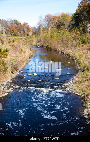 Kleine Stromschnellen in einem Bach, der sich durch eine herbstliche Landschaft mit klarem blauem Himmel schlängelt. Stockfoto