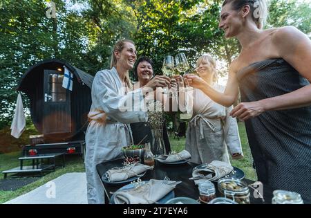Frauen in Bademänteln, die mit Weißwein an einem Tisch mit Geschirr und Servietten vor einer heißen finnischen Sauna tosten Stockfoto