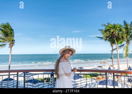 Frau in weißem Kleid und Strohhut auf dem Balkon mit Blick auf den tropischen Strand Stockfoto
