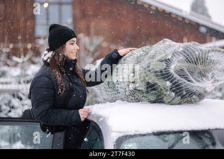 Schöne Frau, die einen Weihnachtsbaum auf das Dach des Autos legt, um ihn nach Hause zu bringen. Lebende Tannenlieferung Stockfoto
