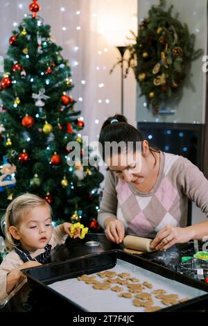 Die kleine Tochter schaut sich die Kekse auf dem Tablett an, während Mama den Teig mit einer Nudelnadel ausrollt Stockfoto