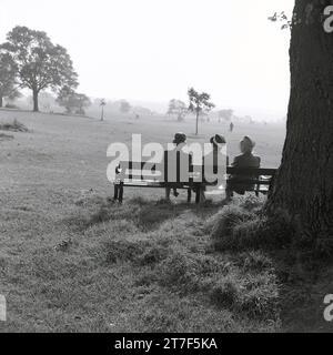 1960er Jahre, historisch, drei ältere Damen in Mänteln und Hüten sitzen nebeneinander auf einer Bank in einem Park, mit Blick auf die umliegende Landschaft, England, Großbritannien. Stockfoto