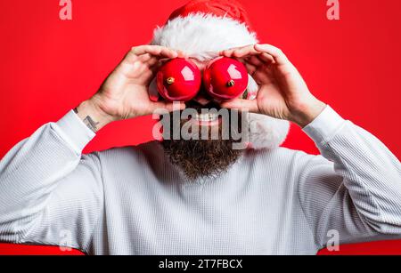 Bärtiger Mann mit Weihnachtsmann-Hut mit zwei Spielzeugen für Weihnachtsbaumaugen. Weihnachtsstimmung. Frohe Ferien. Weihnachtsmann mit dekorativen Weihnachtskugeln in der Nähe Stockfoto