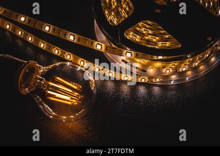 Weicher Fokus der transparenten Glühlampe auf schwarzem Tisch in der Nähe des leuchtend gelben LED-Streifens Stockfoto