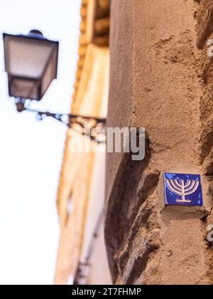 Kleine blaue Fliese mit Menora-Symbol, eingebettet in die Steinfassade im ehemaligen jüdischen Viertel in Toledo, Spanien, Straßenlaterne im alten Stil. Stockfoto