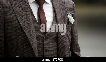 Der Bräutigam trägt einen stilvollen Hochzeitsanzug in braunen Tönen, eine Krawatte und ein Boutonniere im Knopfloch. Elegante Hochzeitsmode. Stockfoto