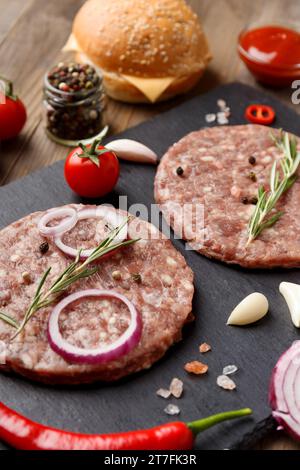 Rohe, frisch gemahlene Rinderburger, Rosmarin, Gemüse und Gewürze auf einer schwarzen Steinplatte auf Holzhintergrund. Zutaten für einen Hamburger. Stockfoto