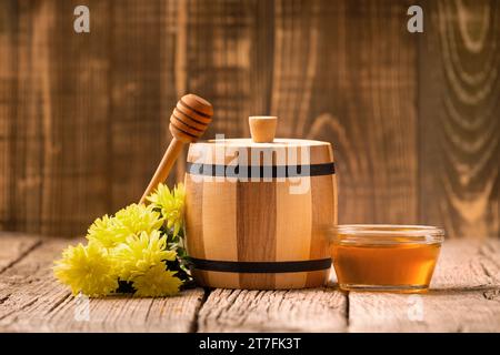 Honig in einem kleinen Holzfass und einer transparenten Glasschüssel, einem Holzdipper, gelben Blumen auf einem gereiften Holzhintergrund, Nahaufnahme. Stockfoto