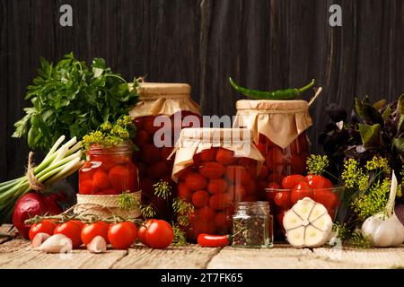 Kirschtomaten in Dosen in geschlossenen Gläsern, Gewürze und Kräuter zur Marinade auf rustikalem Holzgrund. Stockfoto