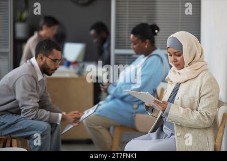 Junge ernsthafte Bewerberin im Hijab, die Fragen des Visumantragsformulars liest, während sie es gegen andere Personen ausfüllt Stockfoto