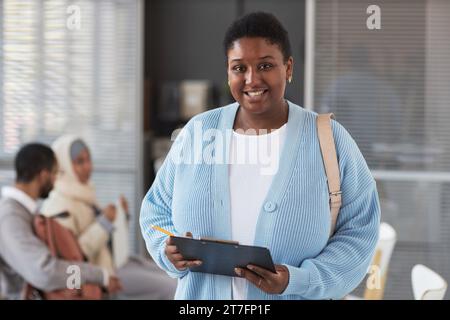 Glückliche junge afroamerikanische Frau in ruhiger Luxuskleidung, die vor der Kamera im Visumantragszentrum steht und das Formular ausfüllt Stockfoto