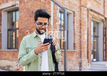 Porträt eines jungen erfolgreichen Studenten außerhalb des Universitätsgeländes, eines Mannes, der lächelt und die App am Telefon benutzt, Nachrichten schreibt und soziale Medien durchsucht, eines indianers, der das Telefon mit dem Rucksack hält. Stockfoto