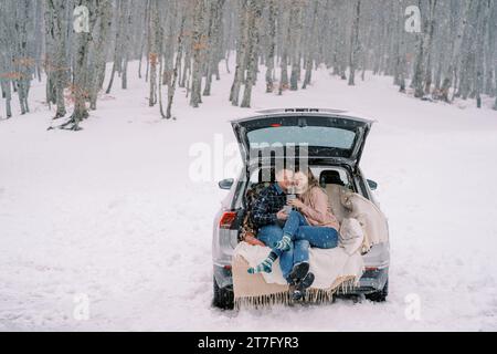 Paare mit Tassen Kaffee sitzt im Kofferraum eines Autos auf Decken unter Schnee im Wald Stockfoto