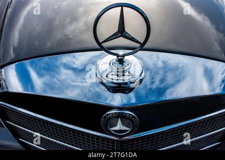 Nahaufnahme der klassischen Mercedes-Benz-Kapuze und des Kühlergrills mit Chrom, die blauen Himmel und Wolken reflektieren und die zeitlose Eleganz des Fahrzeugs symbolisieren. Stockfoto