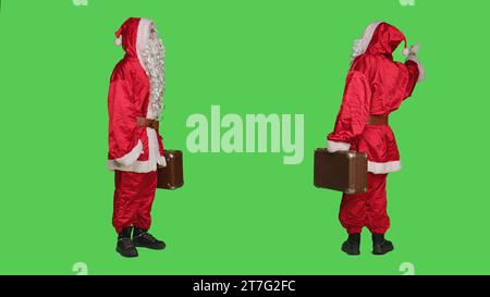 Vater weihnachten trägt Koffer im Studio, trägt traditionelle Festkostüme und roten Hut. Saint-Nick-Charakter mit Aktentasche, die vor grünem Hintergrund posiert, per Anhalter. Stockfoto