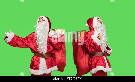 Ein Mann, der sich wie ein weihnachtsmann benimmt, mit einem Sack voller Spielzeug, mit Geschenken und Geschenken über dem grünen Hintergrund. Saint Nick verbreitet weihnachtsstimmung in den Winterferien, Tasche vor der Kamera tragen. Stockfoto