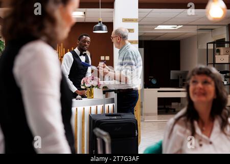 afroamerikanischer Concierge hilft älteren Männern im Ruhestand in der Hotellobby und sorgt für einen reibungslosen Check-in. Ein männlicher Rezeptionist bietet pensionierten Senioren höfliche Hilfe, während er das Reservierungsformular unterschreibt. Stockfoto
