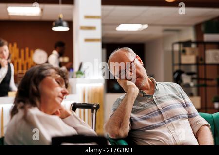 Während ihres Urlaubs entspannen sich abgenutzte Senioren auf einer bequemen Couch im Loungebereich. Ein älteres Paar entspannt sich auf einem Sofa im gehobenen Hotelfoyer. Stockfoto