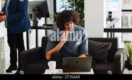 Zahnentzündung in der Betriebspraxis. Mitarbeiter des Unternehmens leiden an Zahnschmerzen, Hohlrauminfektion am Arbeitsplatz. Mann aus dem Nahen Osten mit Karies, der sich im Startup-Freiraum unwohl fühlt. Stockfoto