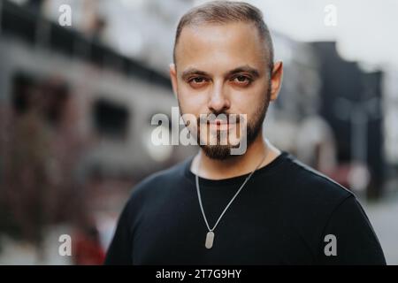 Ein selbstbewusster, attraktiver junger Mann, der draußen posiert. Er lächelt und blickt in die Kamera und steht stilvoll mit einem erfolgreichen Auftreten. Stockfoto