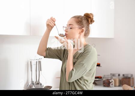 Hübsche junge Frau, die Essen von der Kelle in einer leichten Küche probiert Stockfoto