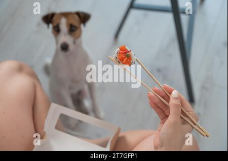 Eine Frau sitzt auf dem Sofa und isst Brötchen. Jack Russell Terrier Hund sitzt auf dem Boden und bittet um Essen von seinem Besitzer. Stockfoto