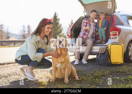 Glückliche Frau, die Hund streichelt, Vater und Tochter sitzen draußen im Kofferraum. Familienreisen mit Haustier Stockfoto