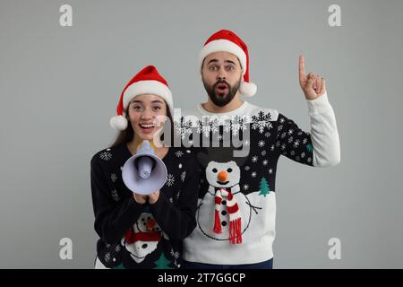 Junges Paar in Weihnachtspullover und Weihnachtsmannmützen, die in Megaphon auf grauem Hintergrund schreien Stockfoto