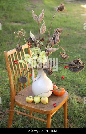 Komposition mit schönen Blumen, trockenen Sonnenblumen und Äpfeln auf Holzstuhl im Freien. Herbstatmosphäre Stockfoto