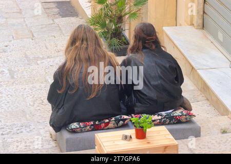 Zwei junge Frauen entspannen gerne auf Kissen auf der Kalksteintreppe, die den Triq San Gwann hinunterführt, in Richtung des Grand Harbour von Valletta. Stockfoto