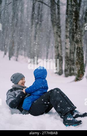 Lächelnde Mutter lag auf dem Rücken im Schnee und ein kleines Kind sitzt auf dem Bauch Stockfoto